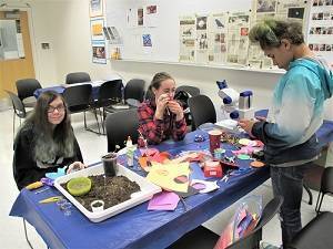 Two teens sit at a table and a third teen stands in front of it. On the table are a microscope, a tray of soil, and pieces of construction paper.